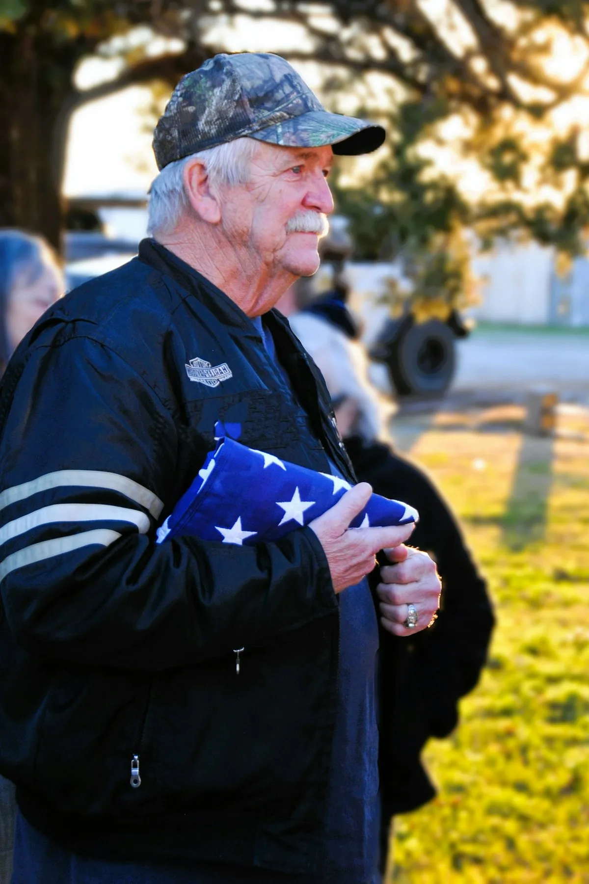 Older veteran in a camo cap and dark jacket holding a folded American flag outdoors